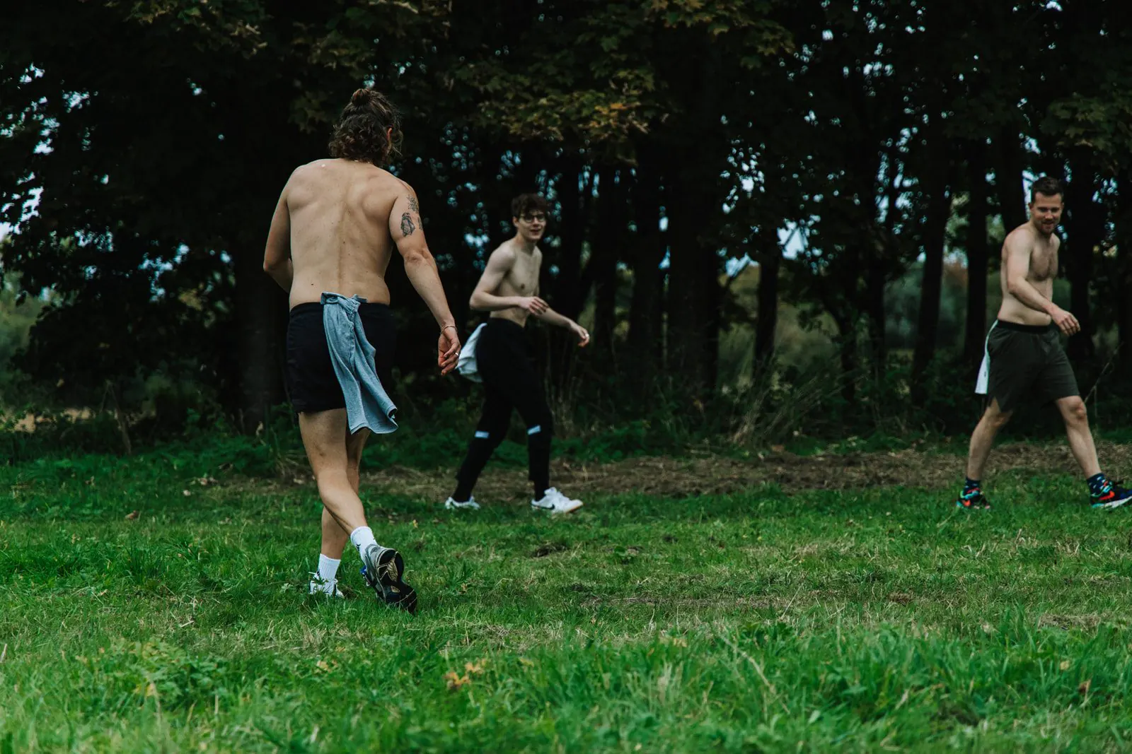 Drie mannen met ontbloot bovenlijf lopen over een grasveld aan de rand van een bos