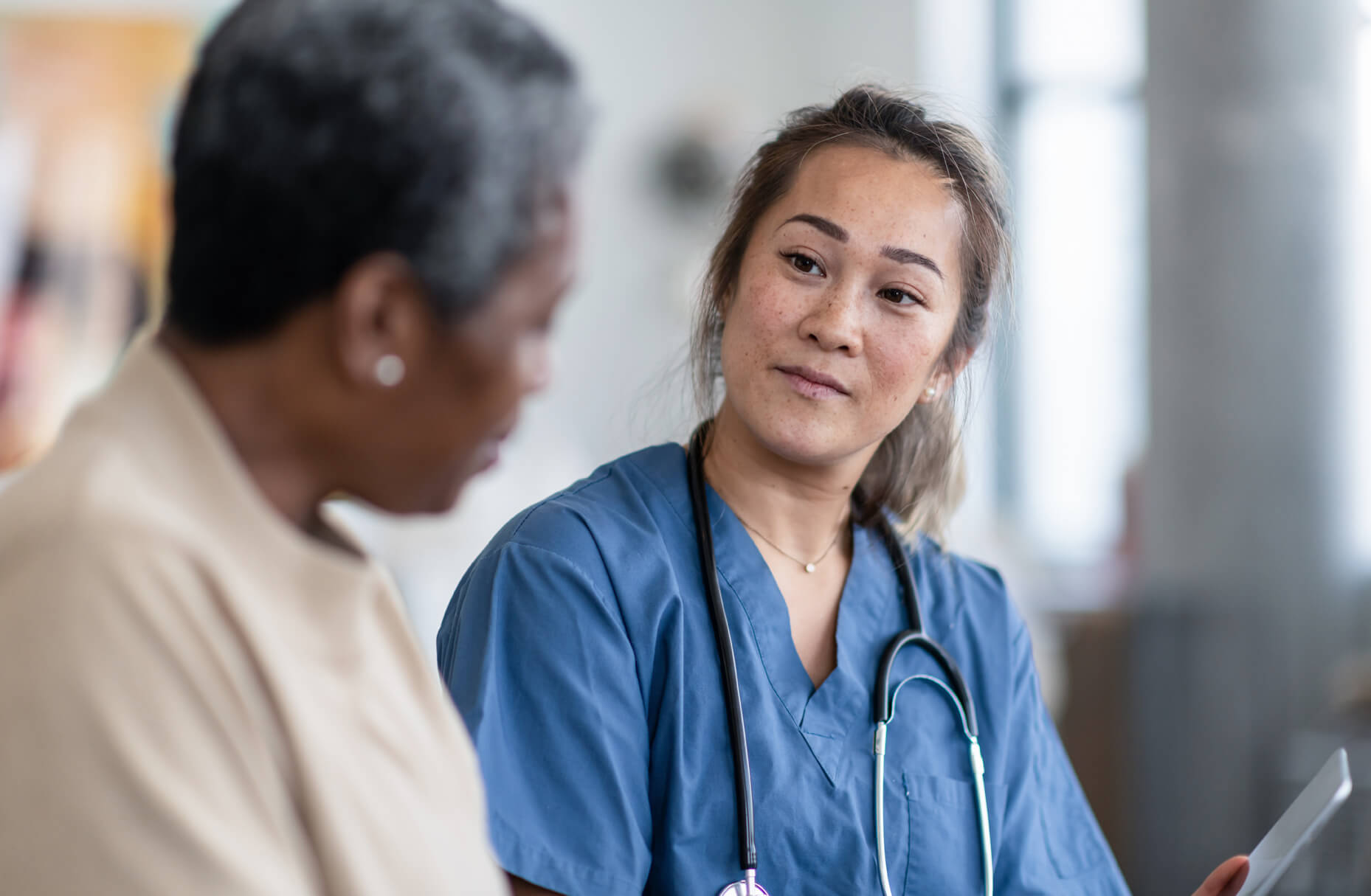 Healthcare professional in blue scrubs with stethoscope listening attentively to patient during medical consultation in bright clinical setting.