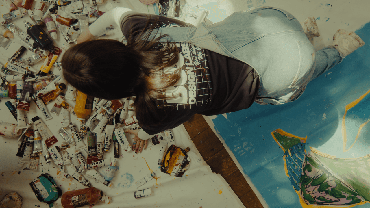 An artist seated on the floor, painting on a large canvas while surrounded by brushes and scattered paint