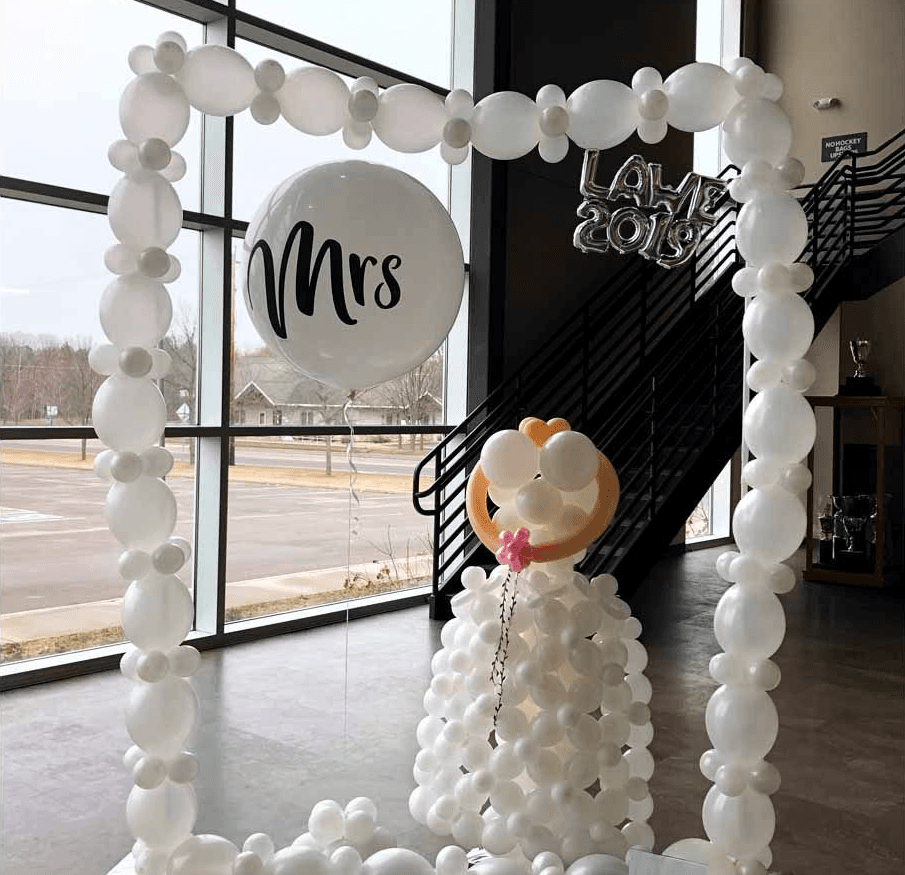 A creative display of white balloons forms a framed backdrop with a large balloon labeled "Mrs" and additional silver balloons spelling "LOVE 2019" in a modern indoor setting near large windows and a staircase.