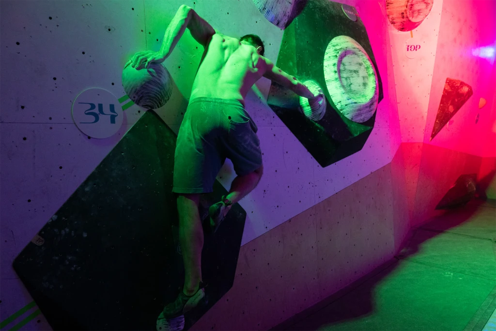 Mężczyzna wspinający się na ścianie wspinaczkowej z uchwytami, podświetlonej zielonym i różowym światłem.