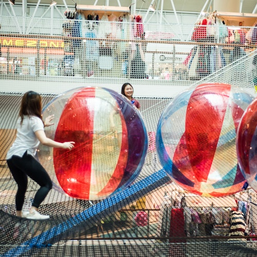 Women playing with large, colorful inflatable balls on netted play structure in a mall, with stores visible in the background.