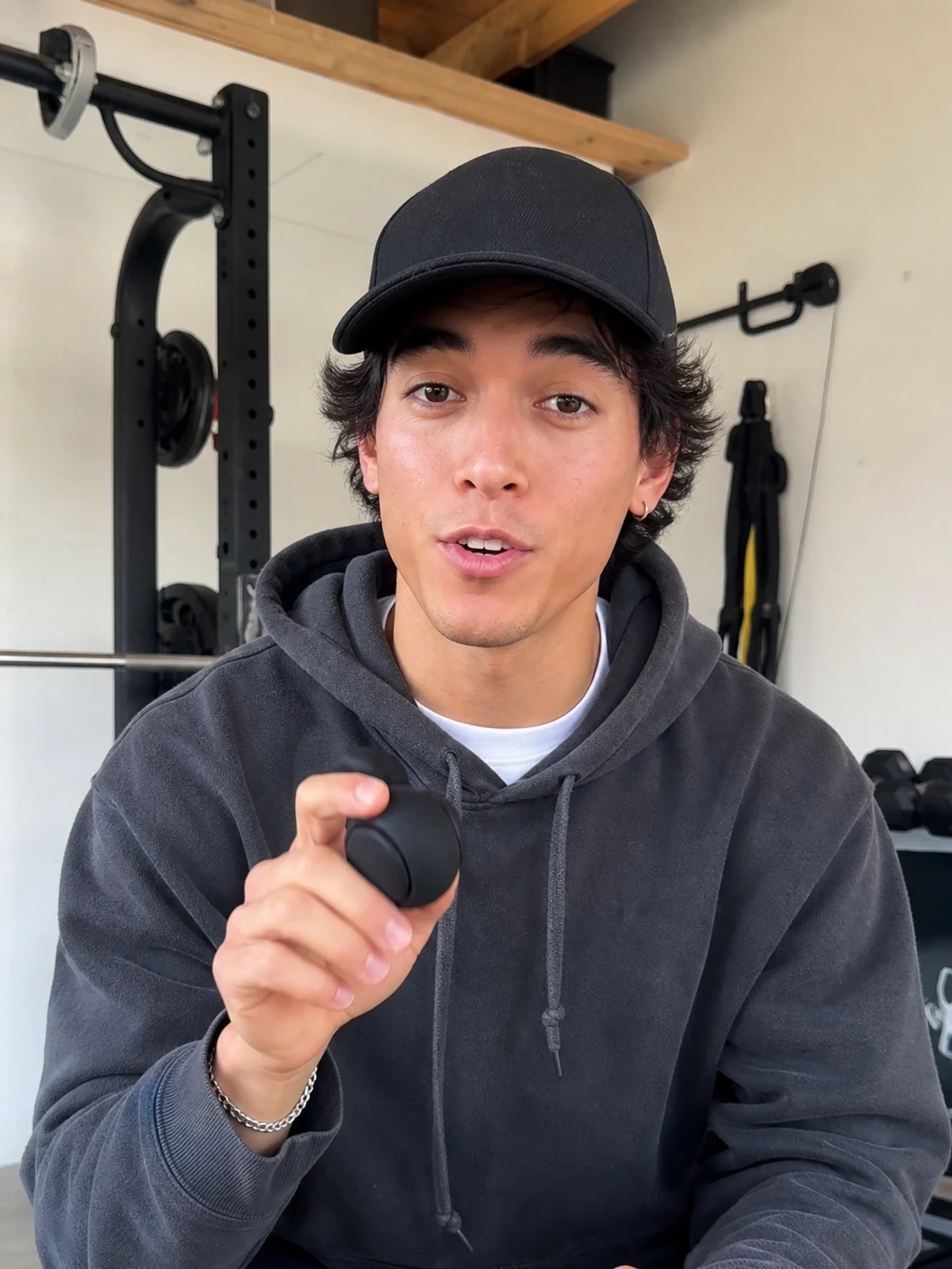 UGC-style photo: streetwear creator, in a small home gym, demonstrating a simple hand-sized product while talking to camera, slightly wider waist-up shot.