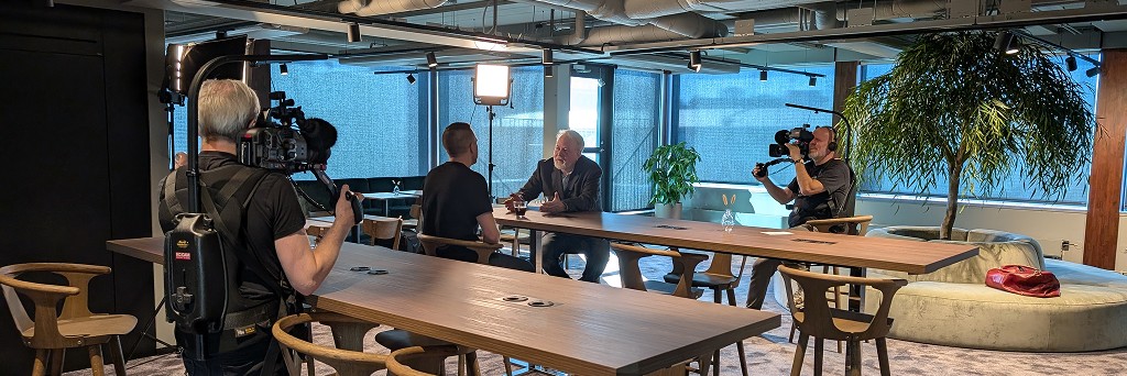 En filminspelning i ett kontorslandskap där två kameramän filmar ett samtal vid ett bord