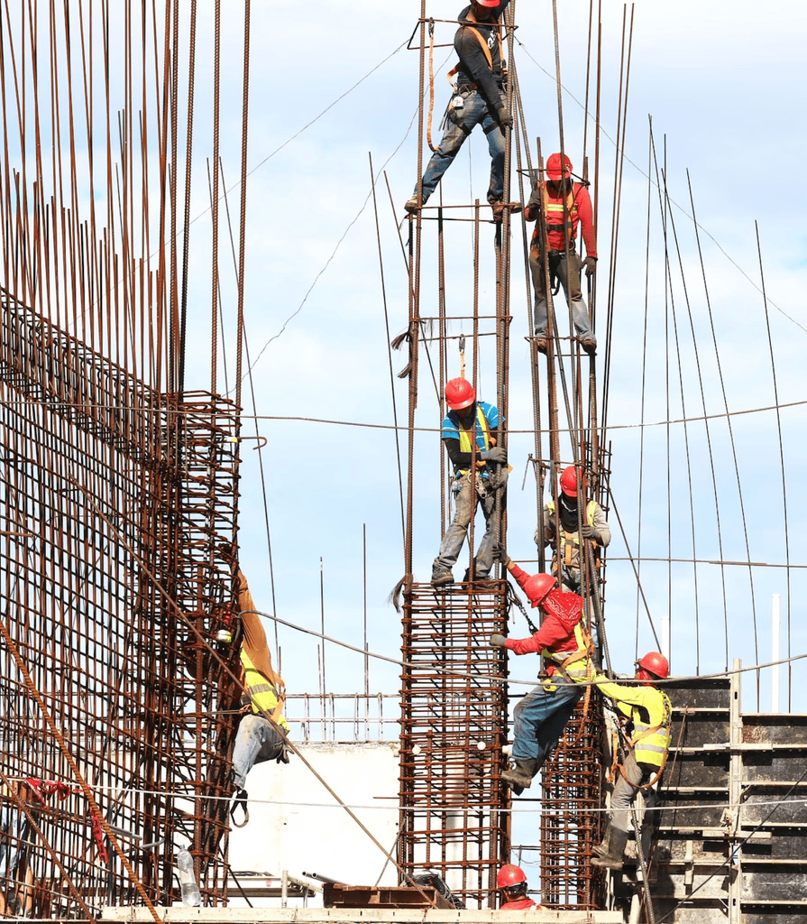 Construction workers in red helmets and safety vests work on tall rebar structures against a blue sky, conveying teamwork and industriousness.