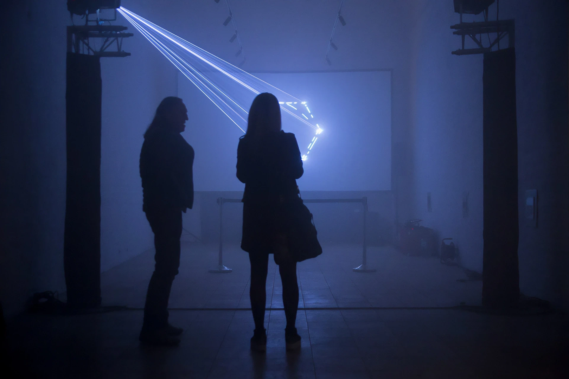 Two individuals watch a laser light show on a screen.