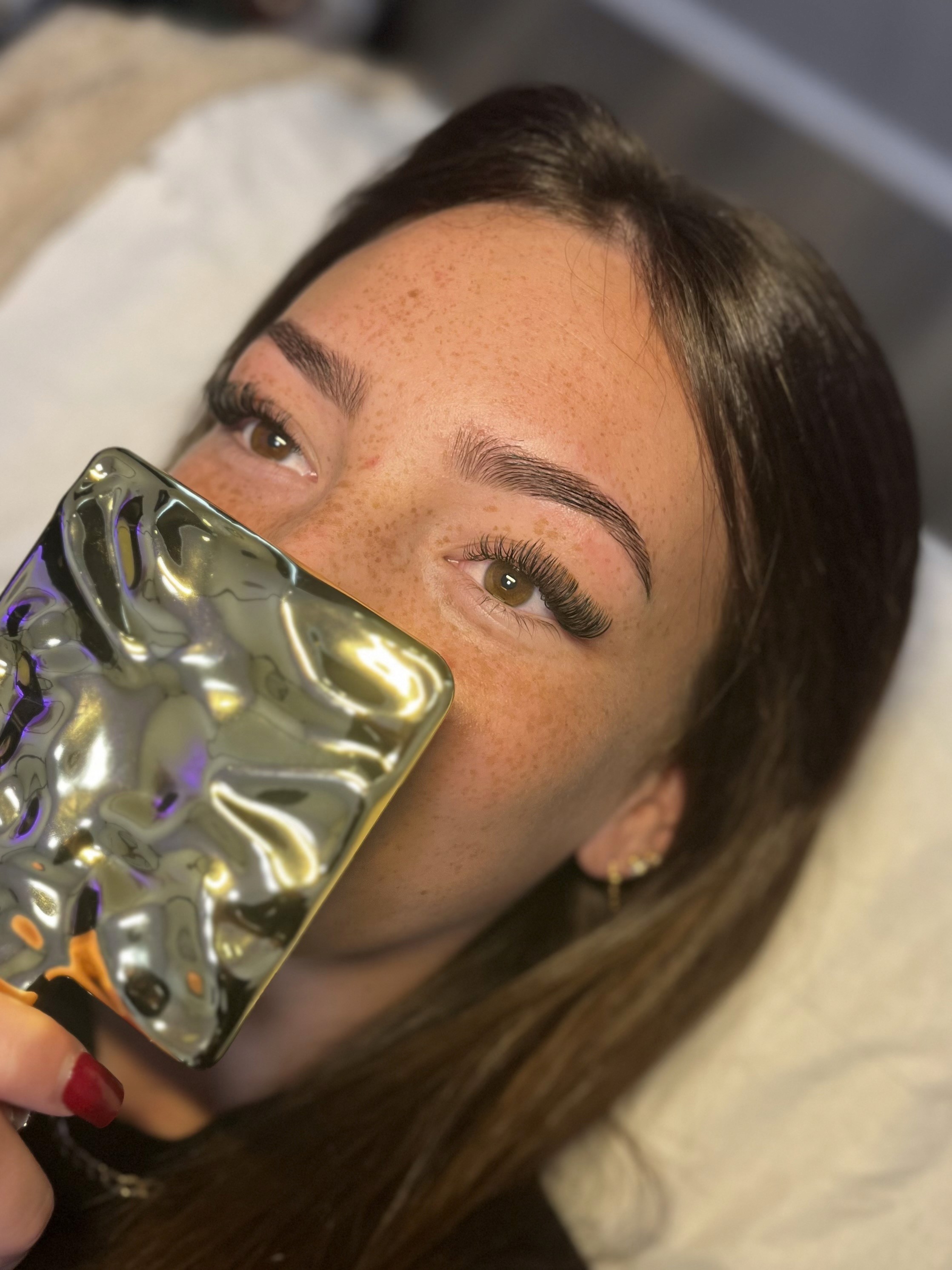 A person holding a phone up to their face, lying on a bed with neutral-colored bedding, and long hair.