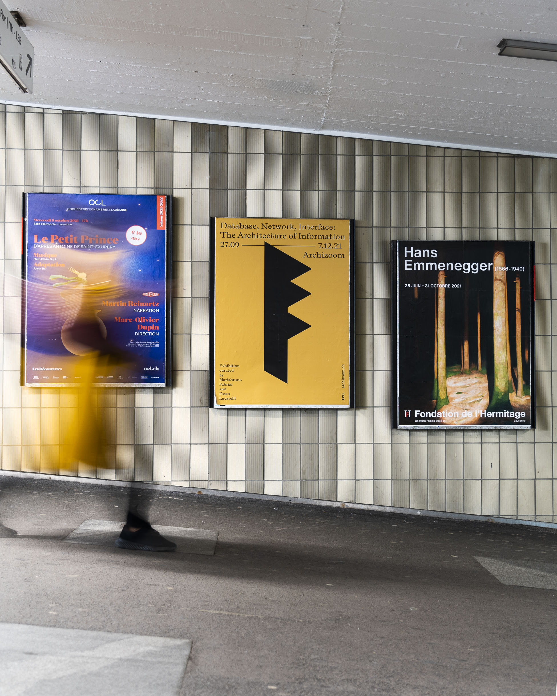 Photo prise dans un passage sous voie montrant une personne passant devant une série d'affiches parmi lesquelles l'affiche Archizoom EPFL