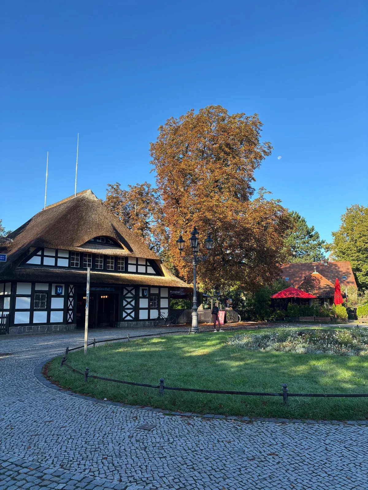 Ein historisches Fachwerkhaus mit traditionellem Reetdach und großem Vorplatz, umgeben von alten Bäumen in Steglitz-Zehlendorf.