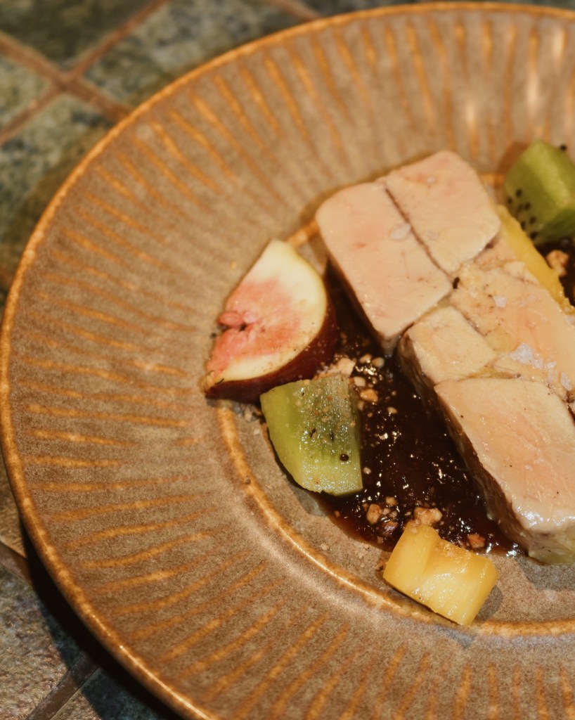 A plate of foie gras terrine served with tropical fruit, including slices of fig and kiwi, drizzled with a rich sauce.