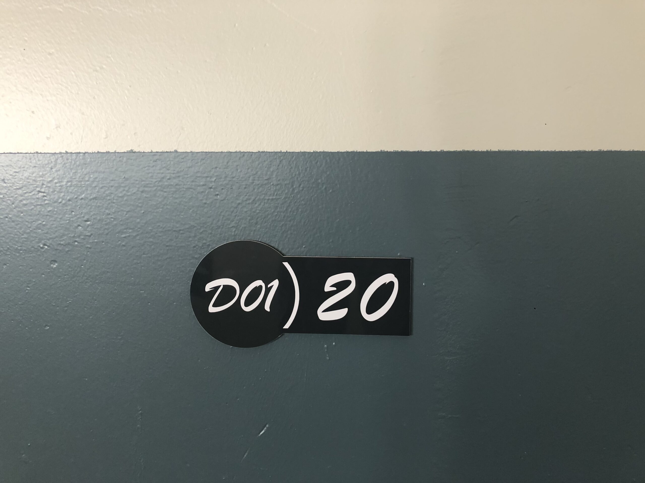 Plaque de porte noire avec inscription blanche « D01 20 » sur un mur bleu sarcelle, au design simple et moderne. L'environnement semble calme et ordonné.
