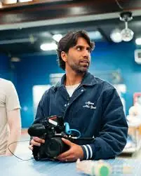 A man wearing a navy blue jacket holds a professional camera in a well-lit studio with blue walls, capturing an attentive expression as he stands beside another person partially visible.