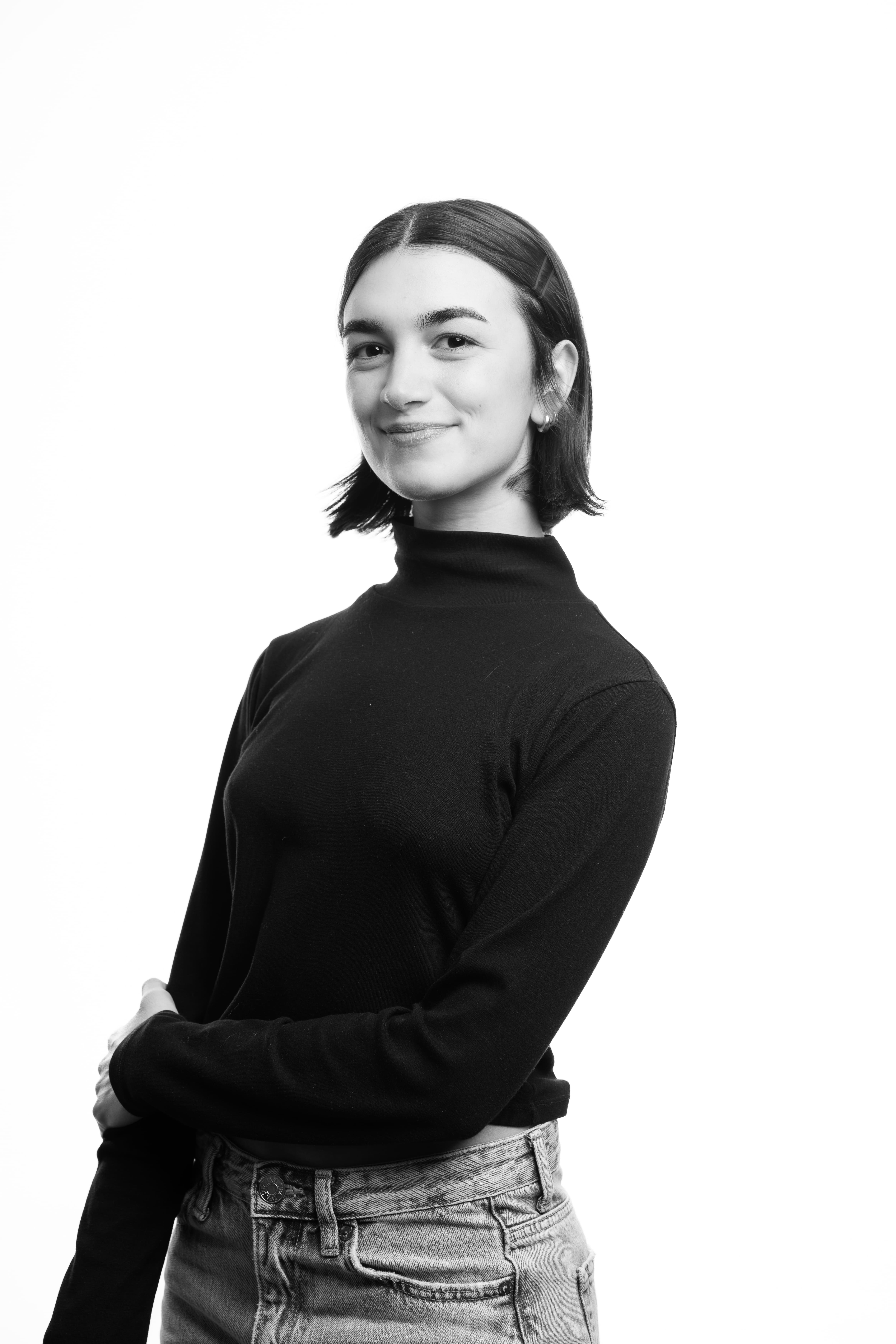 A person with short, dark hair smiles slightly, wearing a black turtleneck and jeans. The black and white image has a confident, calm tone.