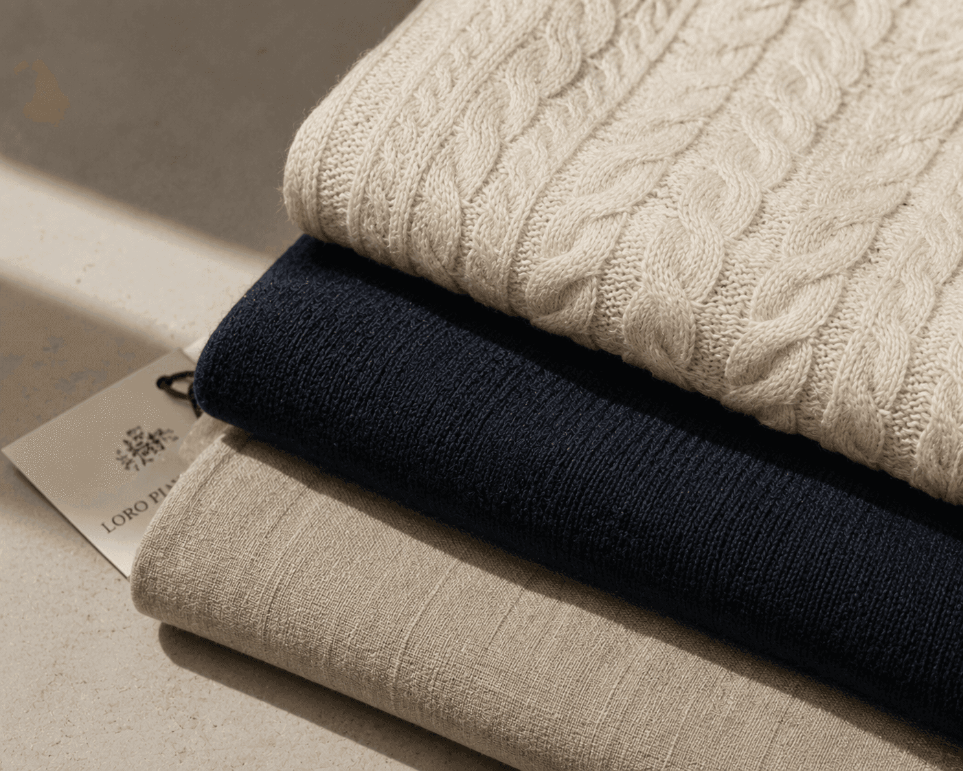 A close-up shot of three neatly folded luxury garments neatly stacked on a light beige stone surface in soft sunlight. The top item is an off-white cream cable-knit wool or cashmere sweater showing intricate rope patterns. Beneath it lies a dark navy fine-gauge knit garment, resting on a beige woven linen or wool fabric. A Loro Piana brand tag is subtly visible, tucked under the left side of the middle navy layer.