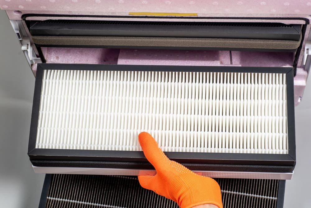 A person wearing orange gloves is installing a white pleated air filter from an air conditioning unit, ensuring optimal indoor air quality in Asheville. The focus is on the filter and the gloved hand, with parts of the air conditioning unit visible in the background.