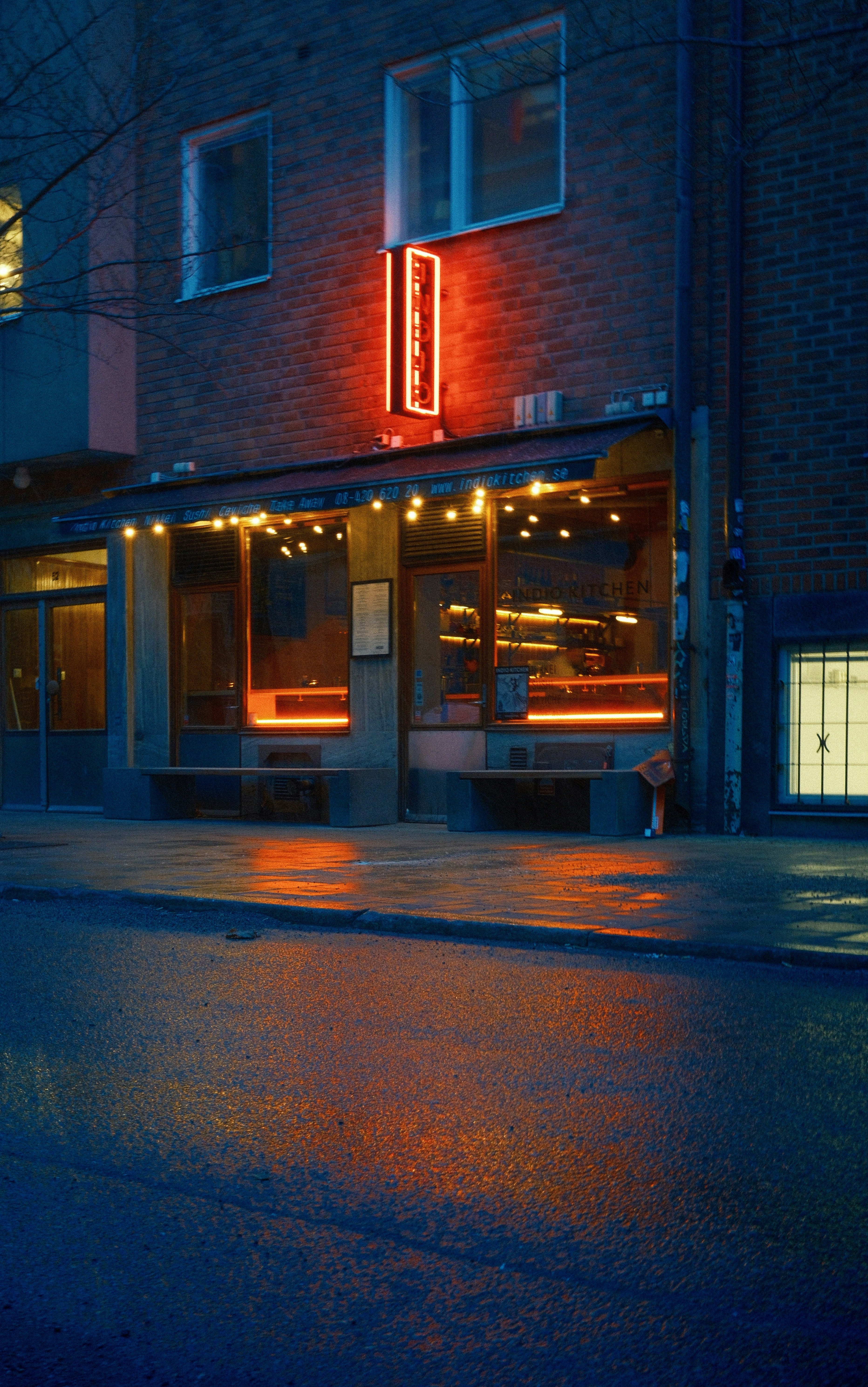 A neon-lit restaurant reflects in wet pavement.