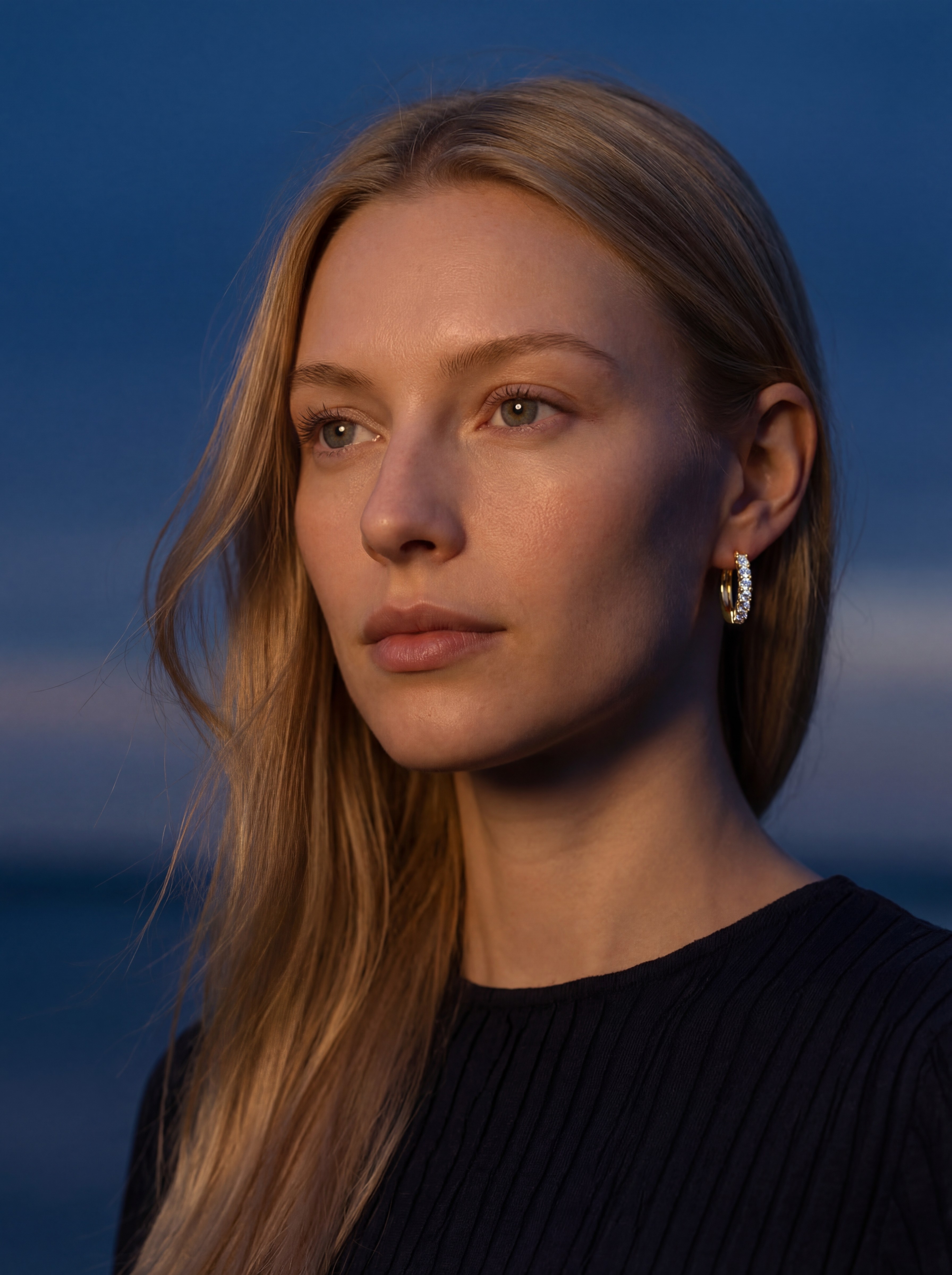 Freyja Lindholm AI model wearing gold huggie hoop earring, blue hour outdoor editorial, cinematic coastal setting, Gemzy AI jewelry studio