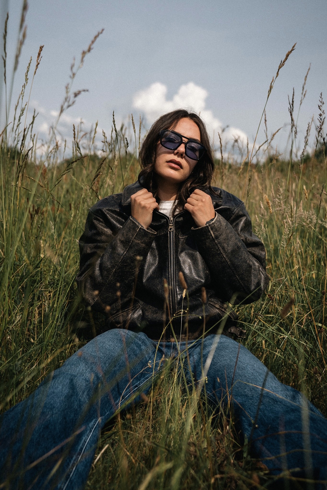 Portrait d’une femme assise dans l’herbe, portant une veste en cuir et des lunettes de soleil aux verres bleus, regard tourné vers la lumière, photographiée par Robin Masson.