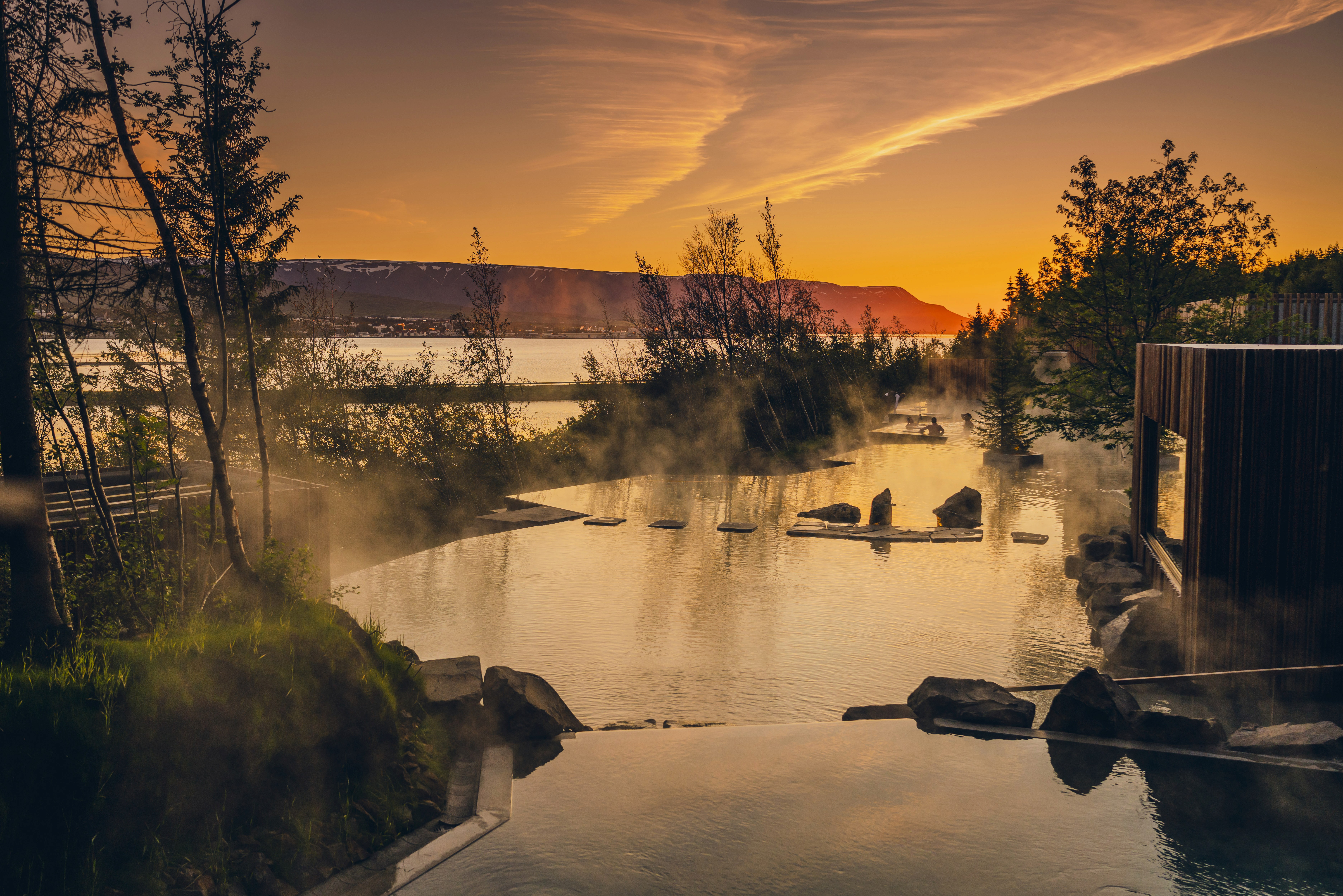View of Forest Lagoon in North Iceland at sunset, with steam rising from geothermal pools nestled among trees and overlooking the fjord, highlighting Iceland’s use of geothermal heat for spa and wellness tourism.