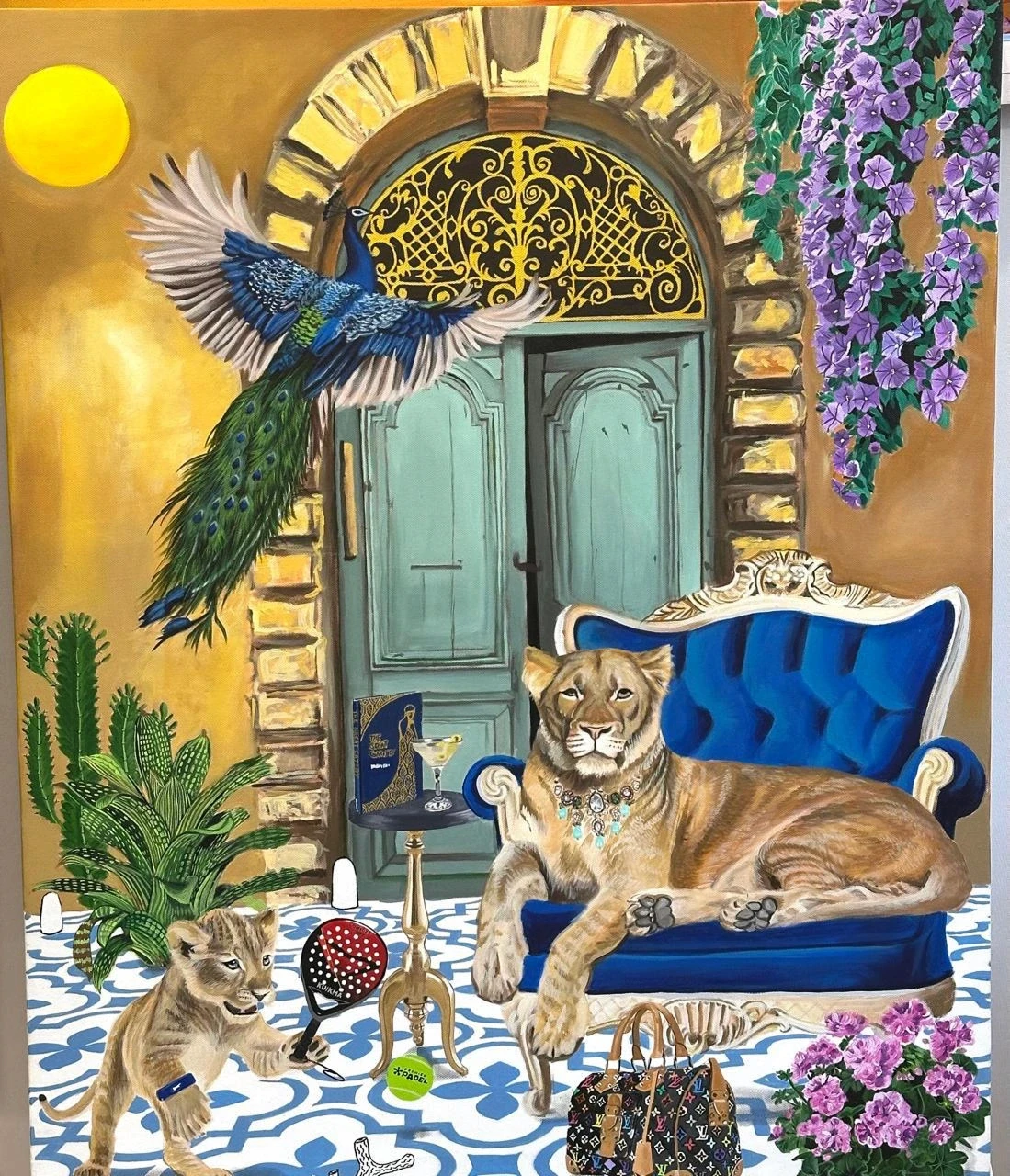 A surreal artwork titled "Belle Époque Lounge" features a glamorous lioness with turquoise jewelry lounging on a blue velvet chair beside a martini glass and vintage book before turquoise doors with golden filigree arches. A playful lion cub swings a paddle with a tennis ball nearby, as a majestic peacock flies above, surrounded by cacti, wisteria vines, patterned tiles, designer bags, and purple flowers under a bright yellow sun.