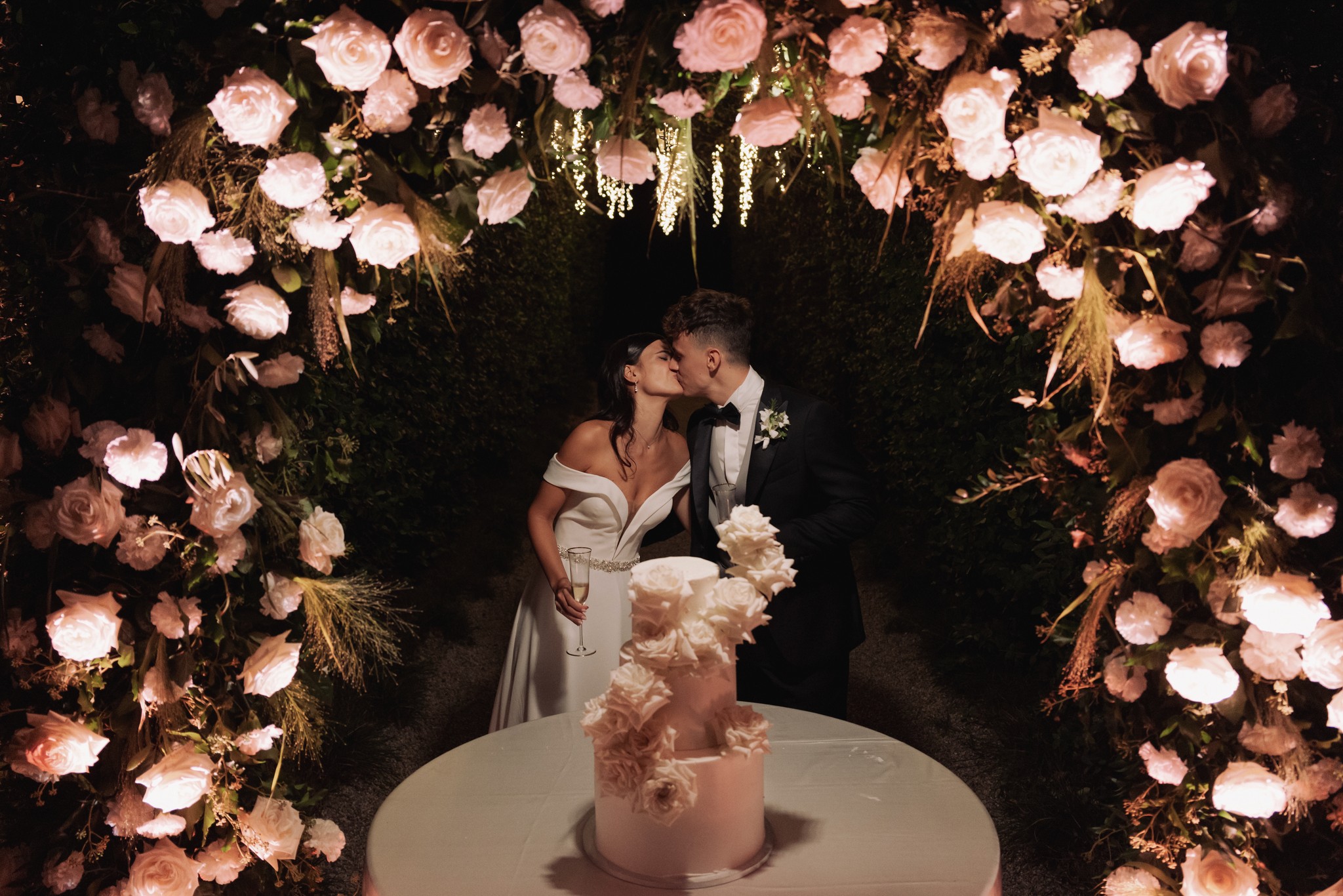 Sposi al taglio della torta sotto arco floreale illuminato di rose rosa e bianche, ricevimento a Villa Negri