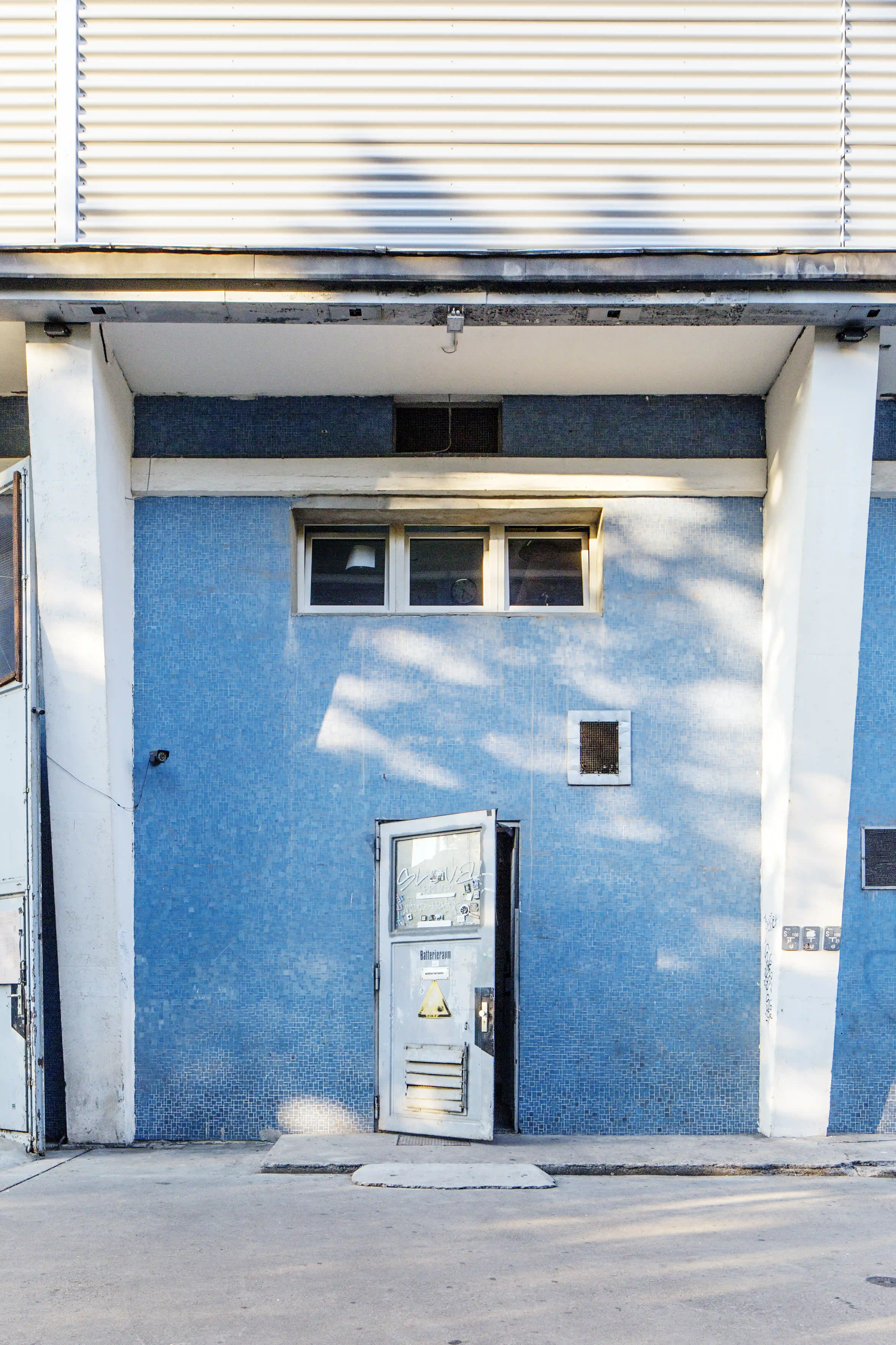 Exterior of OHM CLUB with a blue mosaic-tiled facade, a small white entrance door with window panes and white concrete columns under a corrugated metal band.
