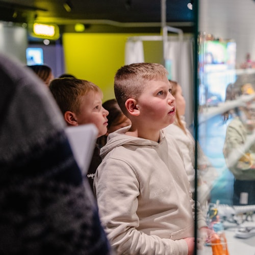 穿着连帽衫的孩子们专注地观看一个室内展览中的展示,周围是人群。