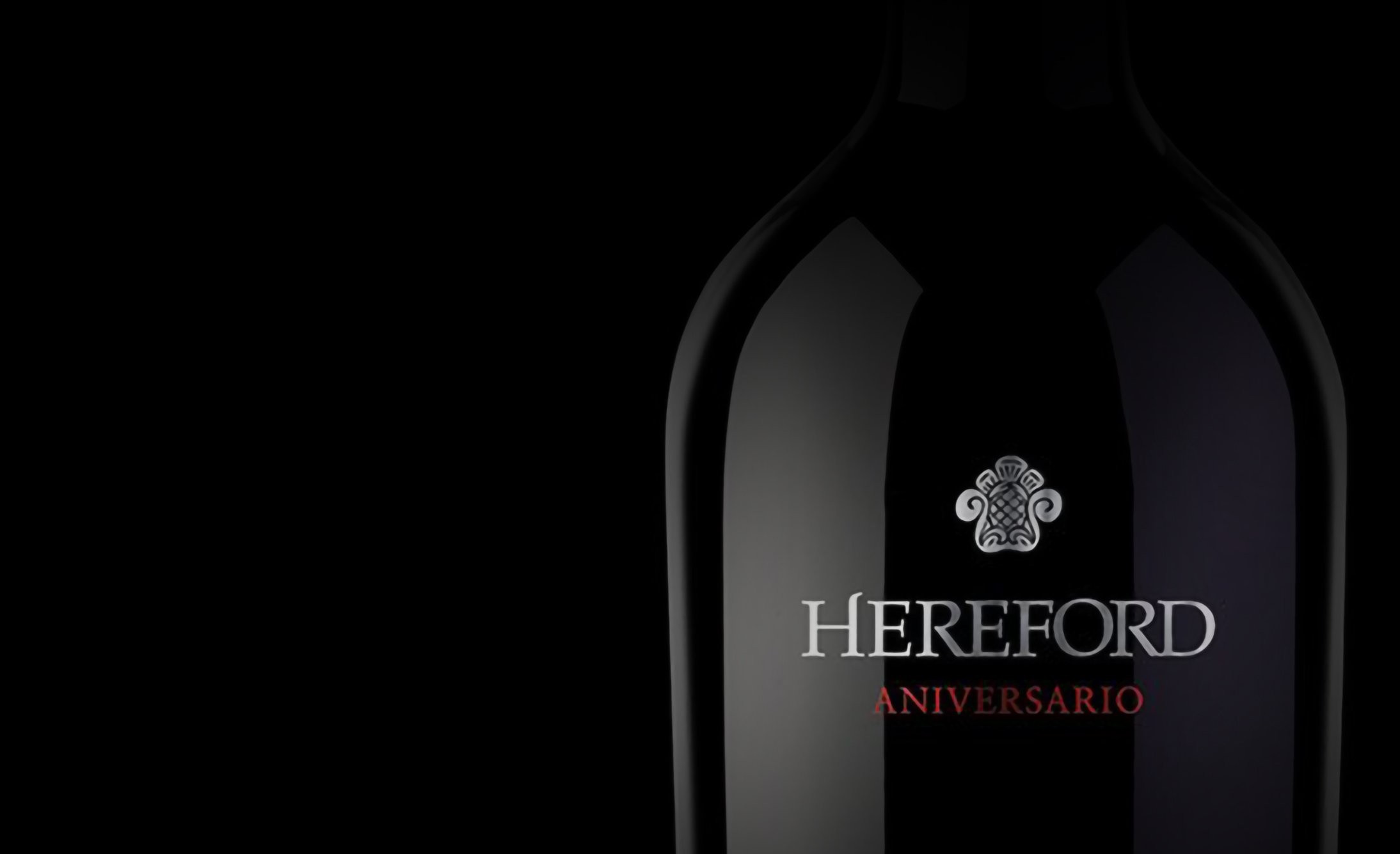 Dark wine bottle against a black background, showing "Hereford Aniversario" text in silver and red with an elegant emblem above. Luxurious and sophisticated mood.