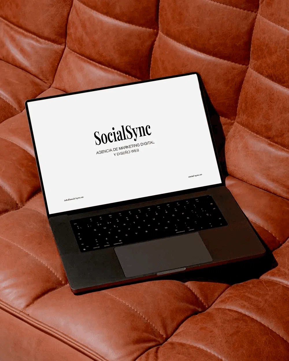 Ordenador portátil sobre un sofá de cuero con las inscripciones «SocialSync» y «Agencia de Marketing Digital».