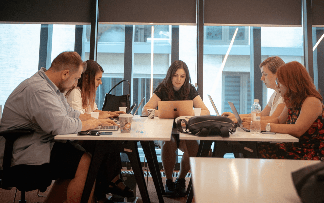 A group of people working together in an office or study environment. They appear to be engaged in collaborative work, with some using laptops and others interacting with each other. The scene conveys a sense of teamwork and productivity.