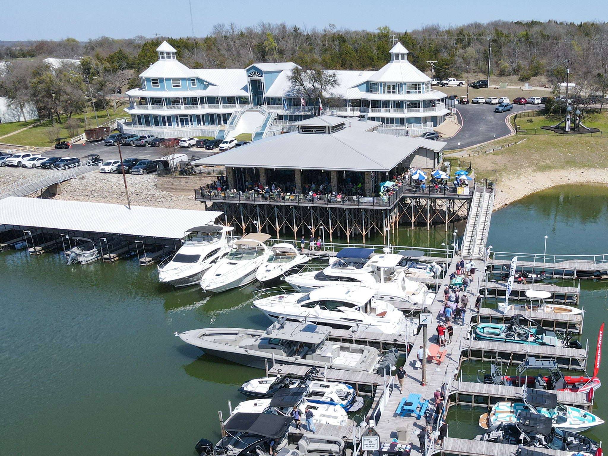 A vibrant marina scene showcases a collection of white yachts and boats docked alongside a bustling pier, with a large, multi-story building featuring unique architectural towers in the background, surrounded by parked cars and lush trees.