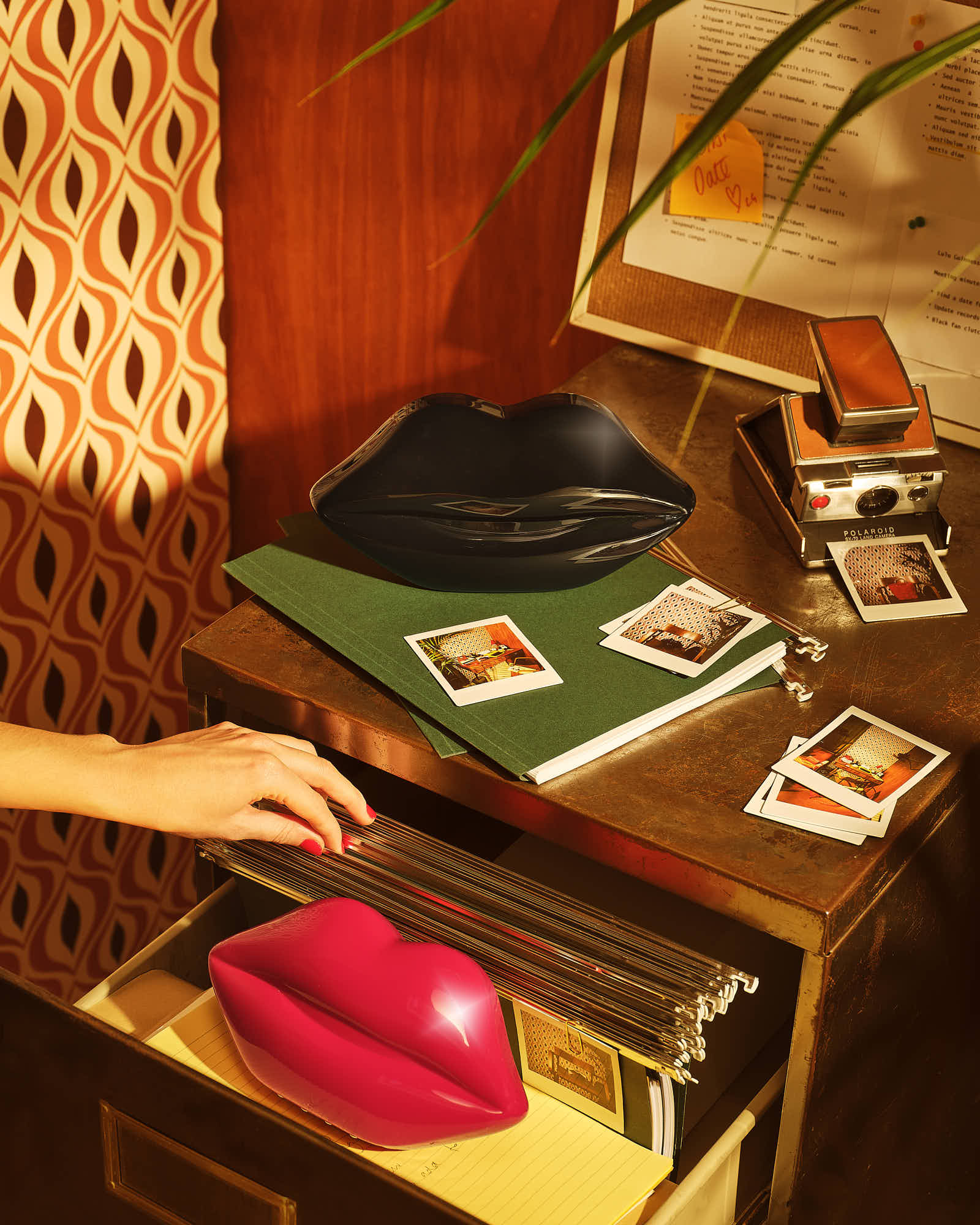 Retro desk scene with Polaroid camera, colorful lips decor, hand organizing files, and patterned wallpaper.