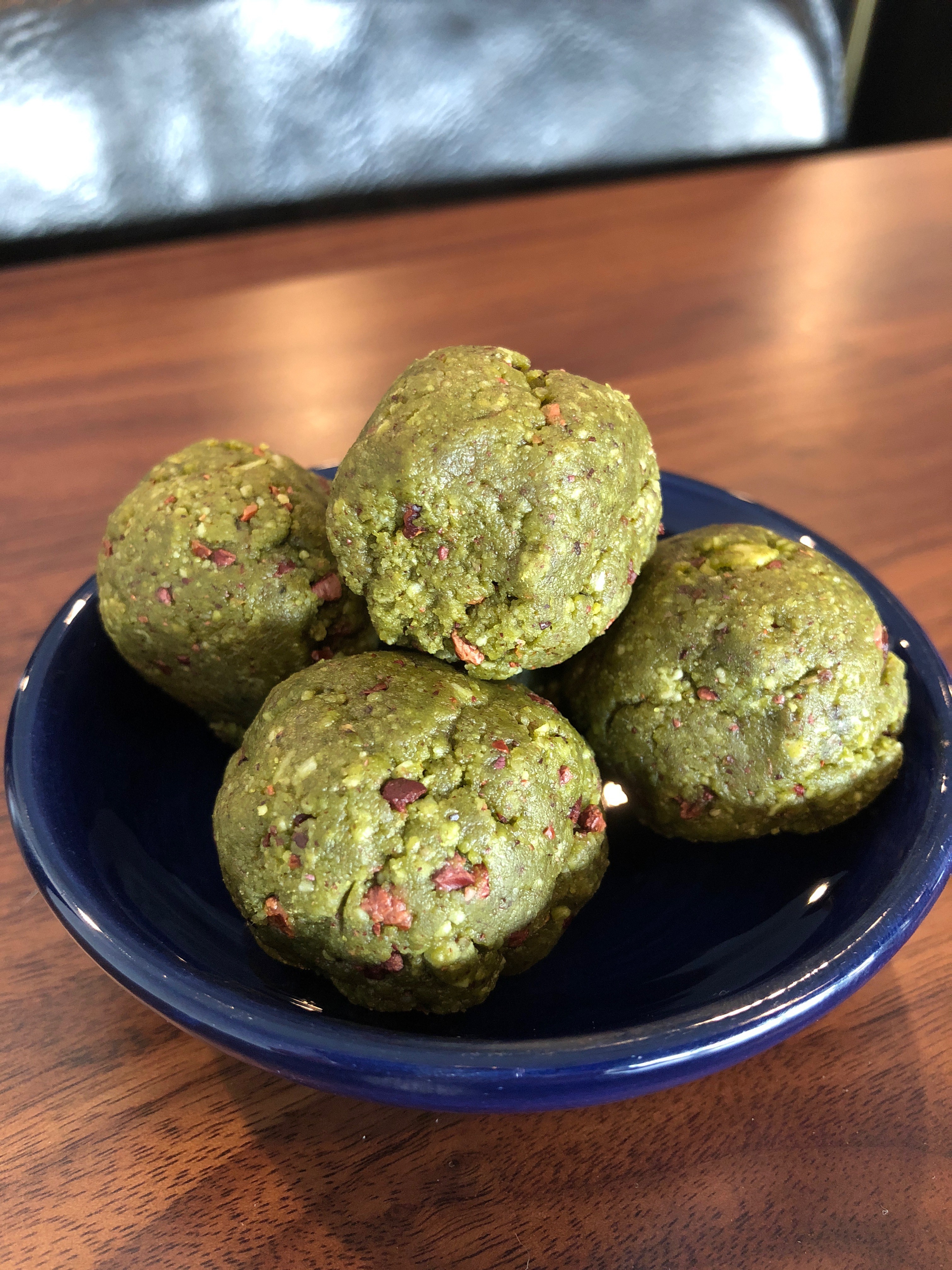A small blue bowl filled with round, green energy bites featuring visible seeds and nuts, placed on a polished wooden table.