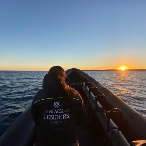 En person med en "Black Tenders Events" jakke sitter foran på en båt ved havet i solnedgangen.