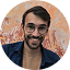 Un homme souriant avec une barbe et des lunettes, portant une chemise bleue ouverte, devant un mur orange texturé.