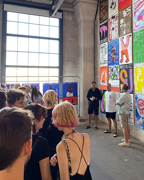 Zdjęcie przedstawiające grupę ludzi, którzy słuchają przemowy otwierającej wystawę artysty oraz organizatorów. W tle znajdują się plakaty artysty.