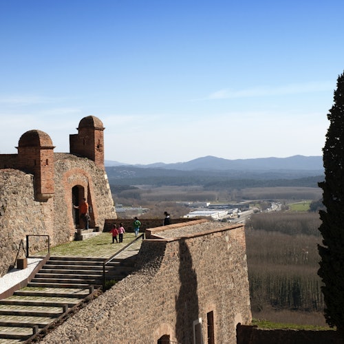 Mga tao na nag-eeksplor ng isang lumang kuta na gawa sa bato na may mga tore, tinatanaw ang malawak na tanawin at mga bundok sa malayo sa ilalim ng malinaw na asul na langit.