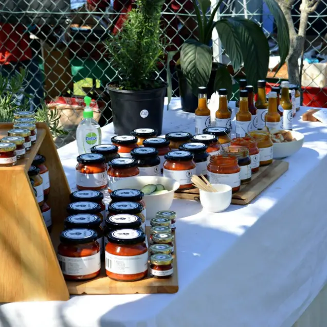 A market stall displays jars of chutney and sauces on a white tablecloth. There are plants, a hand sanitizer bottle, and bowls of sample foods nearby.