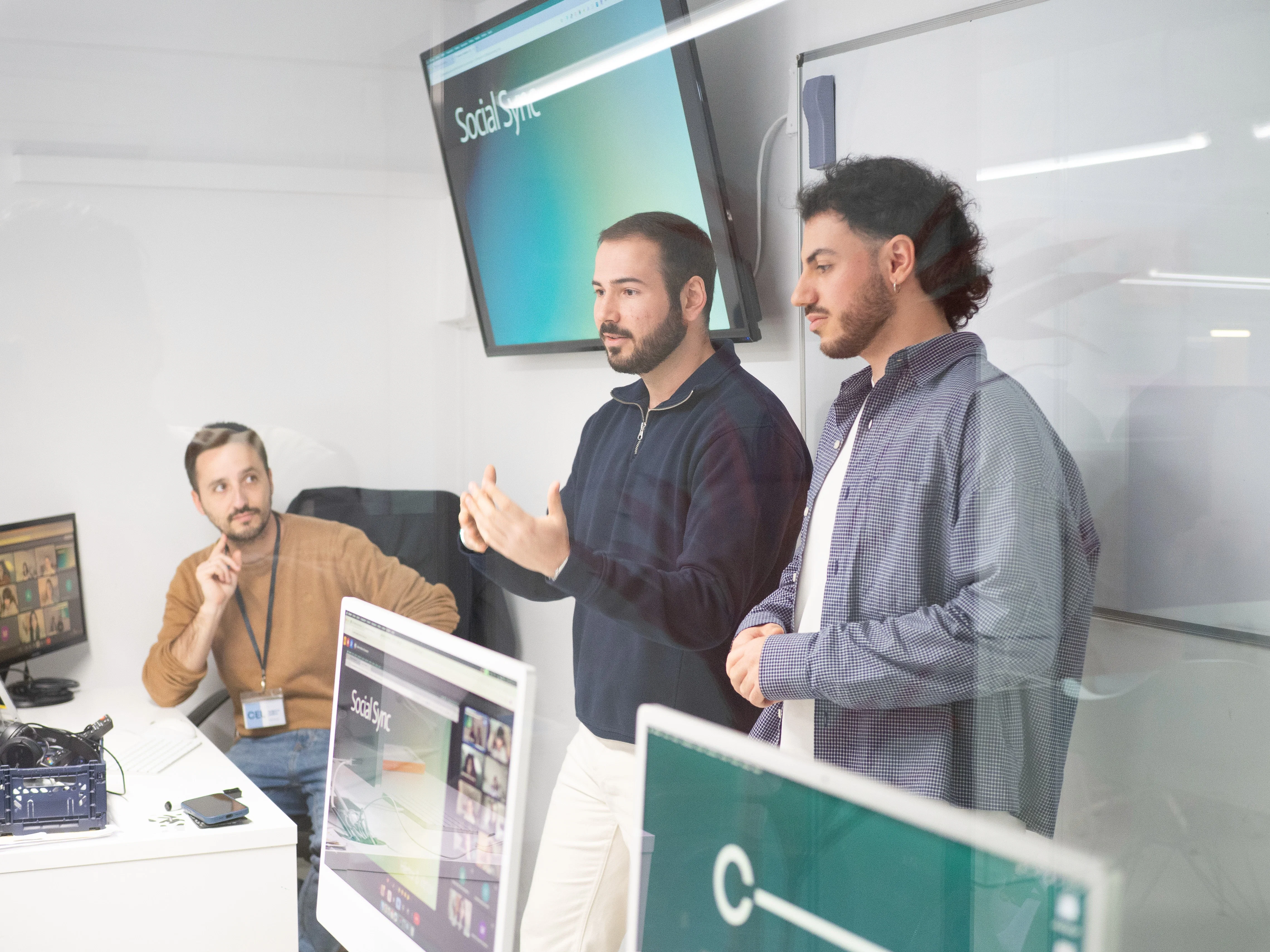 Dos ponentes hablan ante un público, mientras una tercera persona escucha. Aula con pantallas.