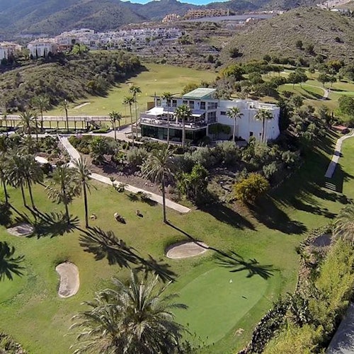 Aerial view of a golf course with a modern building surrounded by trees and hills, with distant buildings and mountains in the background.