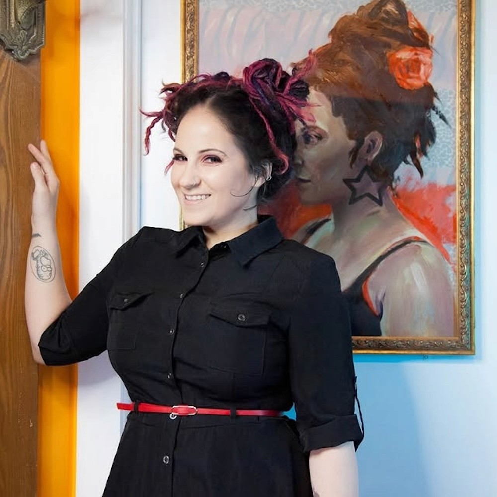 Woman with pink dreadlocks smiles in front of her portrait, wearing a black dress and red belt. The painting behind features a similar pose and colorful background.