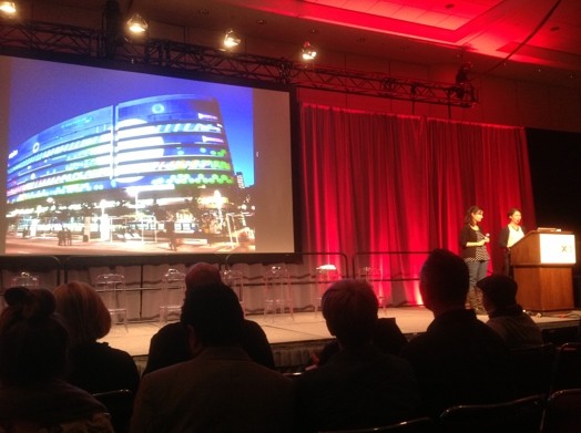 A photo from a hall where two men give a lecture while showing a picture of a building on a screen projector.