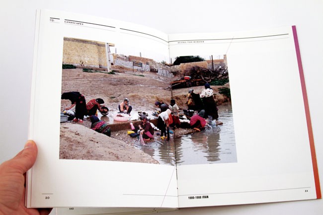 A hand holds open a two-page spread from a book. The left page displays a photograph of people working near a body of water, possibly a river or pond. The right page shows text reading “1980-1988 IRAN.” The pages are white with thin black lines separating sections. The photograph shows individuals engaged in various activities around the water, with some sitting or standing. The background includes a wall or structure. The right page also contains text reading “Along the River.”