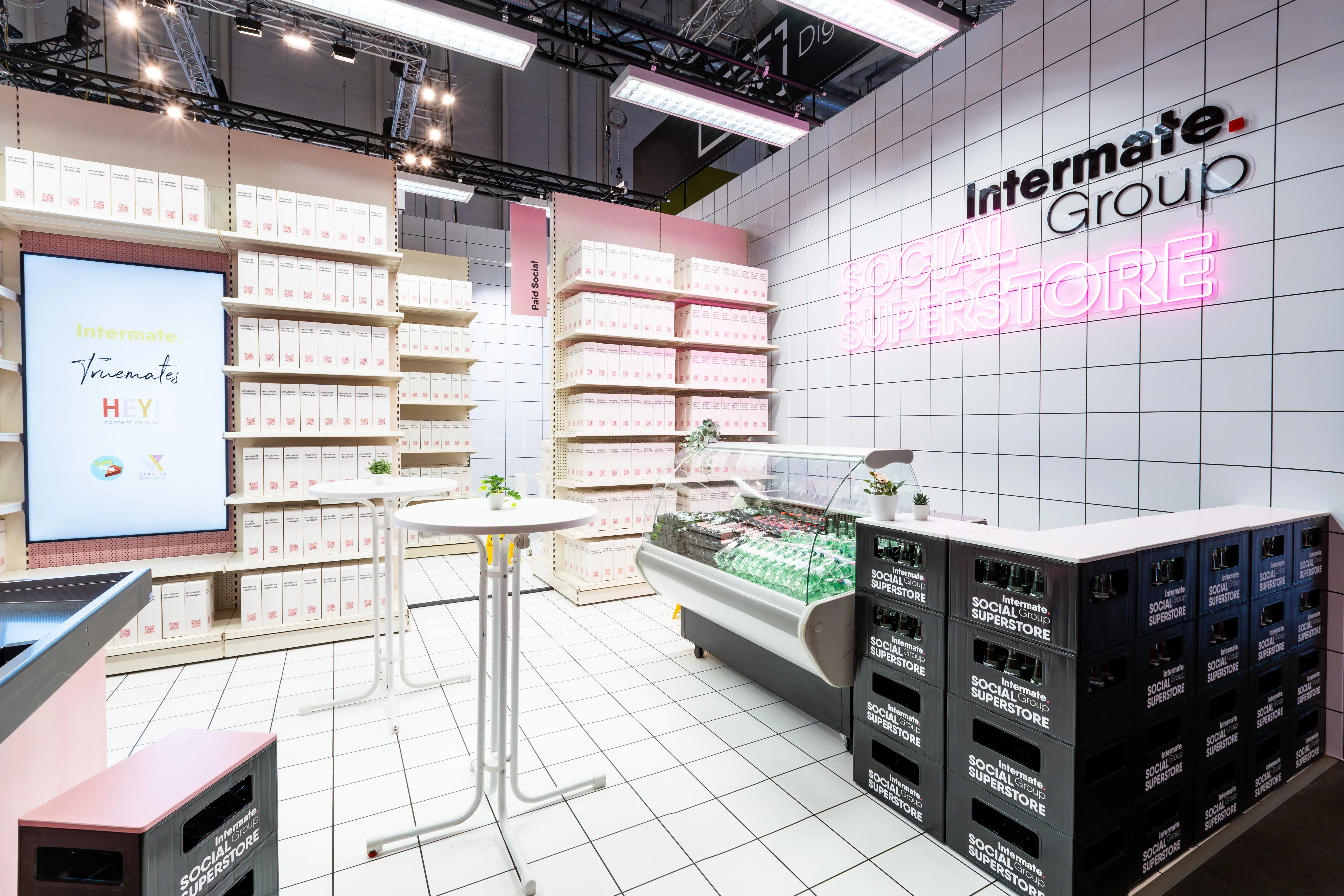 Side view of the Intermate Social Superstore with refrigerated counter, stacked crates, bar tables and pink neon logo.