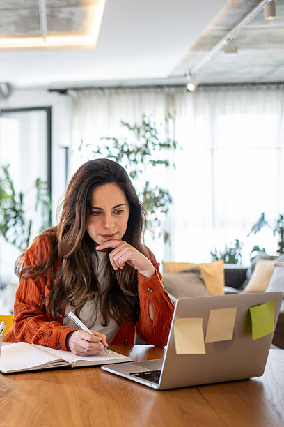 Giovane donna concentrata mentre segue una lezione online, prendendo appunti su un quaderno davanti al laptop con post-it colorati, in un ambiente domestico luminoso.