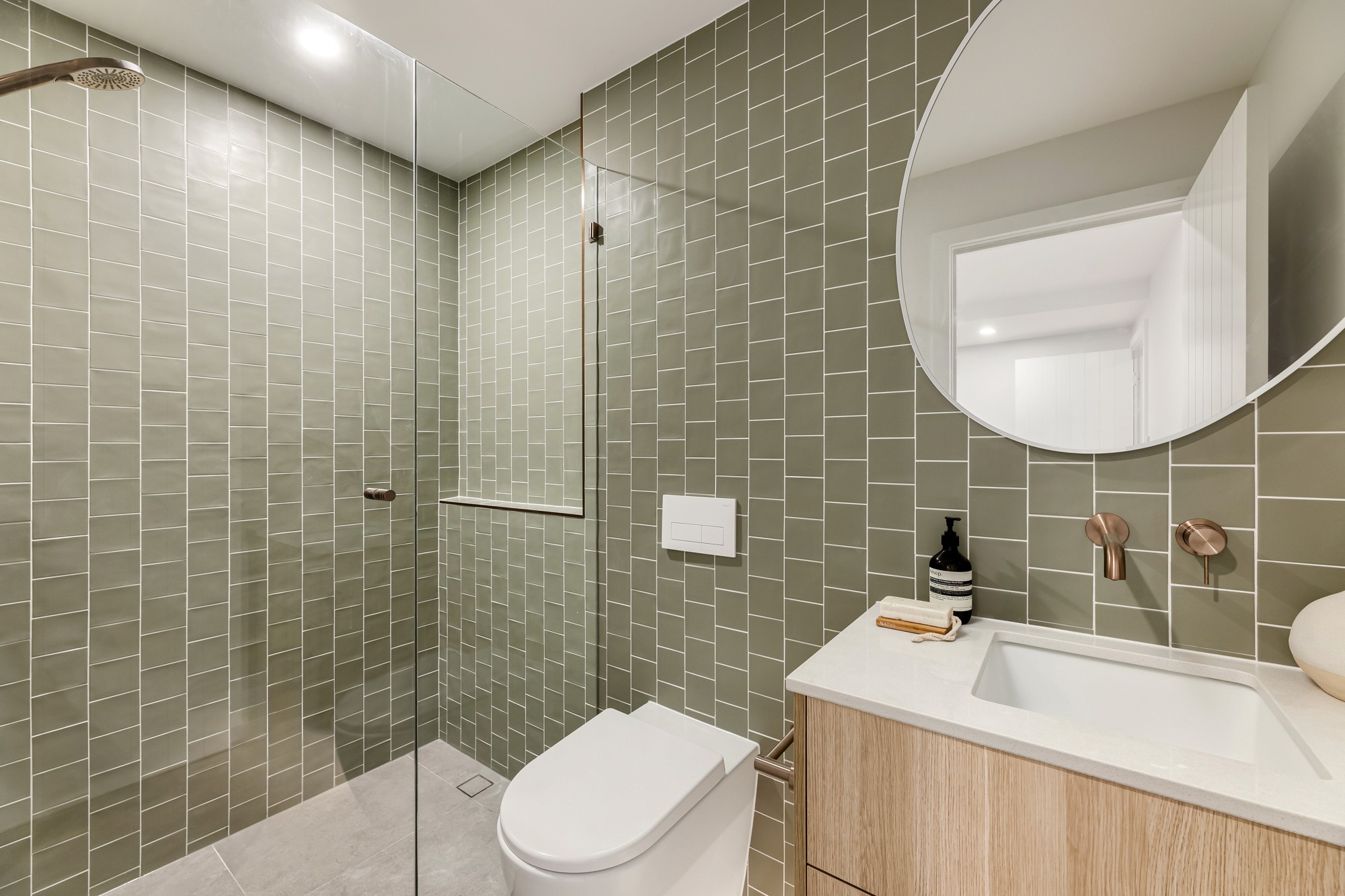 Bathroom with sage tiles, a timber vanity and a round mirror.