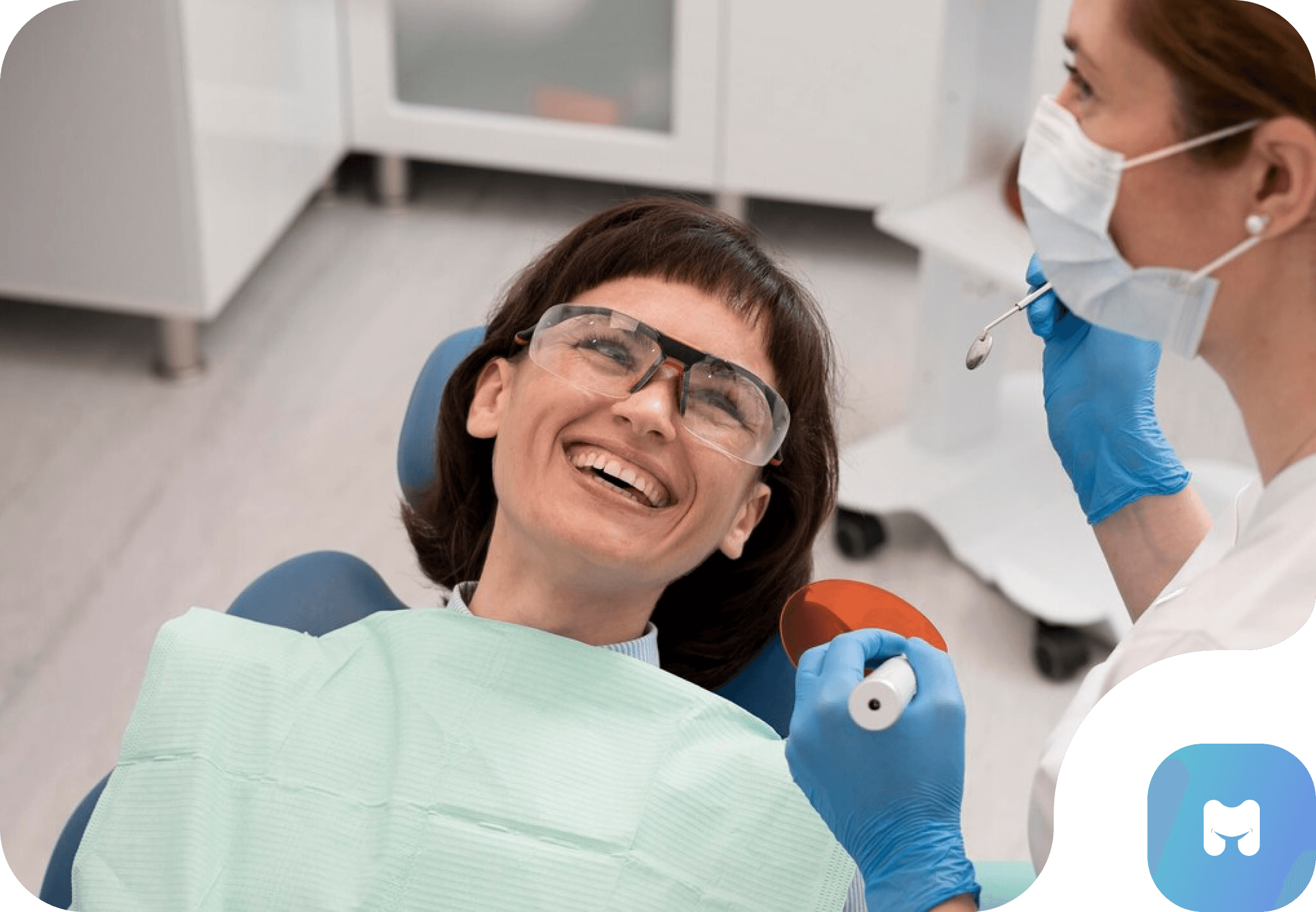 A female patient smiling while sitting in a dental chair, with a dental professional adjusting dental equipment nearby.