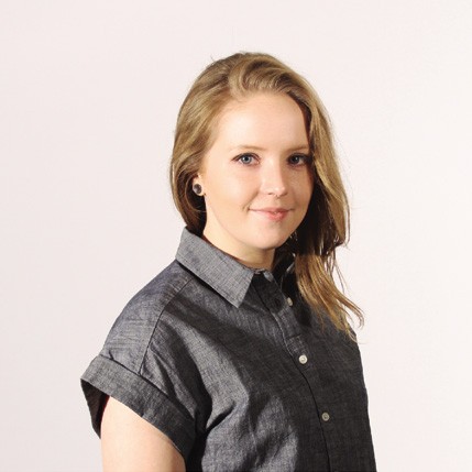 A young woman with shoulder-length light blonde hair wears a dark gray, short-sleeved button-down shirt against a plain off-white background. She is positioned slightly turned to her right, looking directly at the camera with a neutral expression. Her right arm is relaxed at her side, and she wears a small, dark-colored earring in her visible left ear.