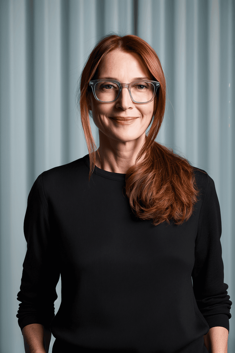 A woman with red hair in a black sweater smiles gently. She wears glasses and stands against a softly blurred curtain backdrop, conveying a calm mood.