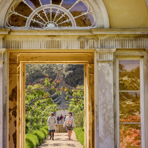 Dvije osobe šeću kroz vrtni prolaz, viđene kroz raskošni kameni dovratnik s ostakljenim gornjim dijelom, okružene drvećem i zelenilom.